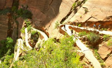 Scenic Zion Canyon trail with hikers and lush greenery on a sunny day.