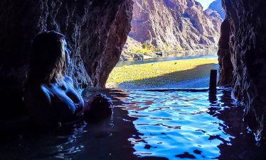Person relaxing in a cave hot spring with mountain view outside.