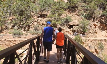 Two hikers on a trail in Zion National Park with rocky cliffs and blue sky.